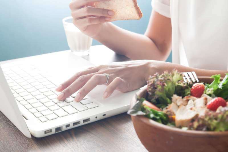 Woman's hand using laptop computer and eating sliced bread with salad bowl in foreground